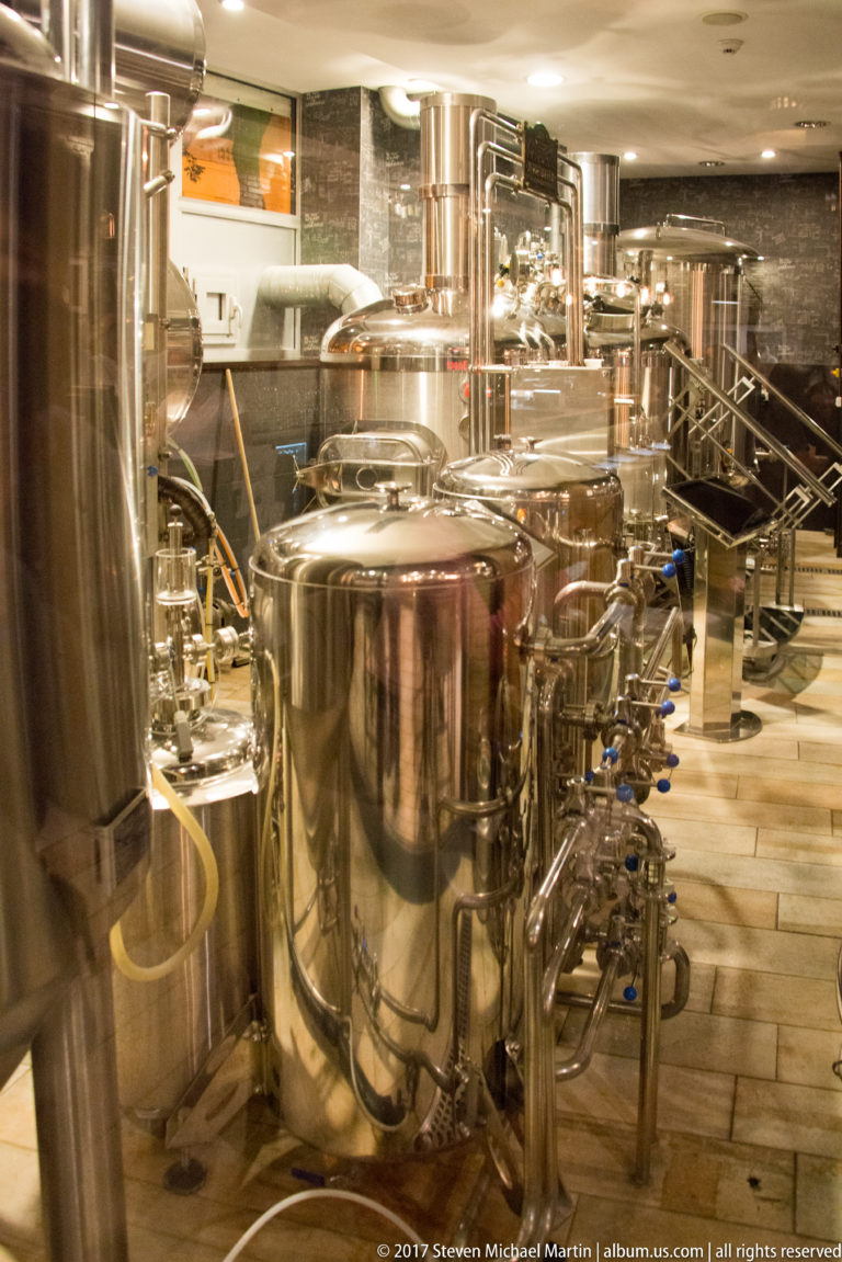 Stainless-steel brewery tanks, pipes, valves, and fittings packed into a bright production room.