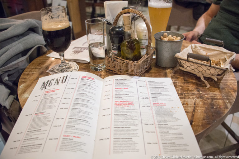 Open restaurant menu on a wooden table with wine, beer, water, bread, condiments, and a cup of snacks.
