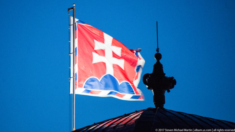 Backlit Slovak flag flying beside the dark silhouette of an ornate rooftop finial against a deep blue sky.