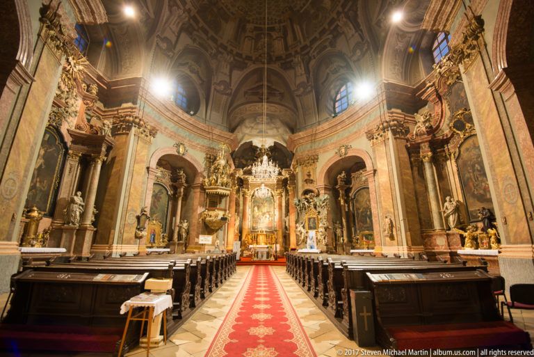 Centered view down the Trinitarian Church nave toward the high altar, with red aisle runner, pews, and ornate Baroque decoration.