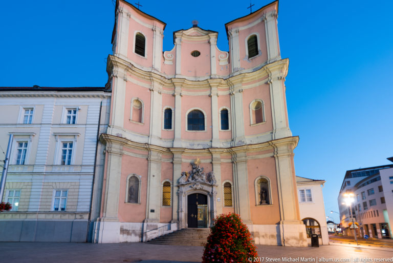 Pink-and-cream Baroque façade of Bratislava’s Trinitarian Church with twin towers and ornate central entrance at dusk.