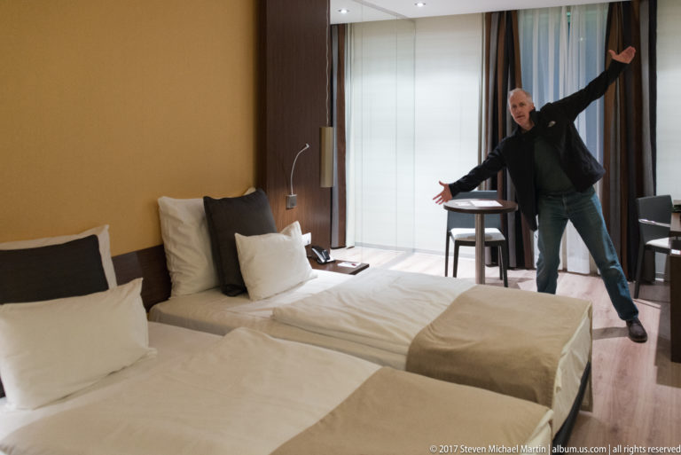 Twin hotel room with two beds, desk furniture, large window, and a person posing near the curtains.
