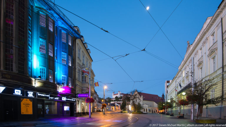 Blue-hour Bratislava street with tram wires, illuminated storefronts, curving tracks, and Bratislava Castle in the distance.