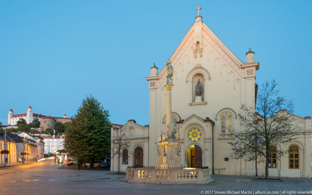 Historic church and devotional column at dusk with Bratislava Castle visible on the hill at far left.