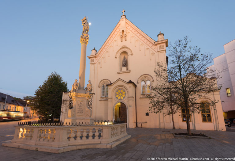 Pale historic church with central gable and saint niche, fronted by a tall devotional column and stone balustrade at dusk.