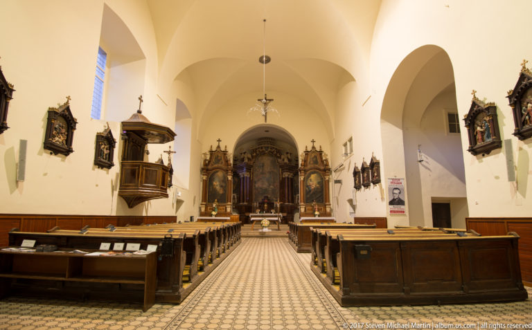 Church nave with dark wooden pews, patterned central aisle, side altars, pulpit, and illuminated sanctuary.