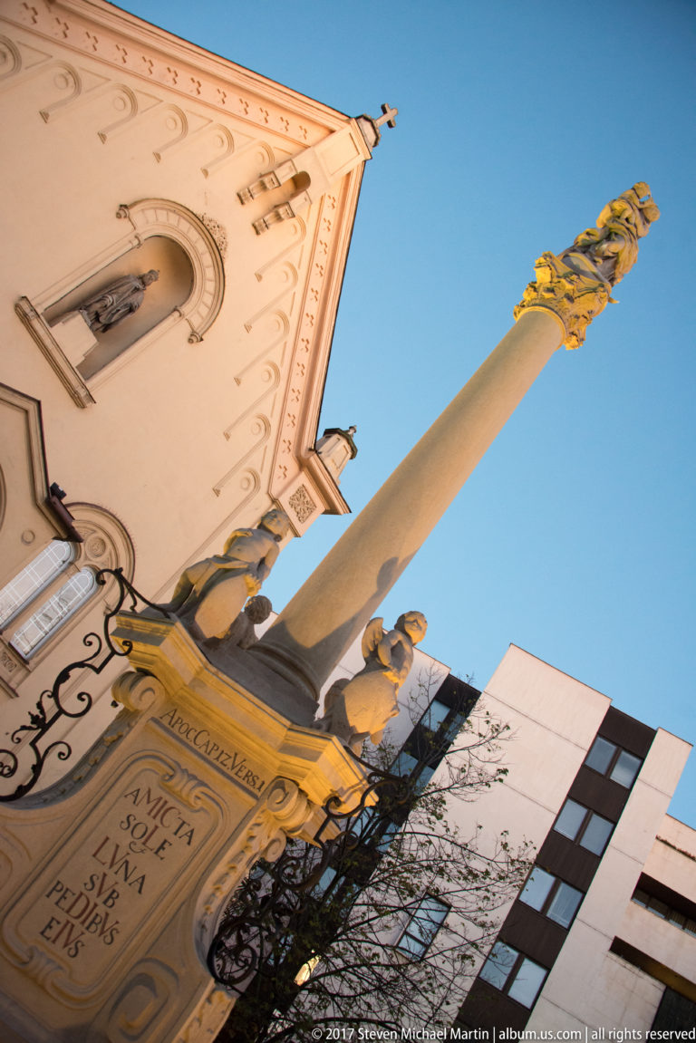 Low-angle view of an inscribed stone devotional column with sculpted figures beside a church façade in Bratislava.