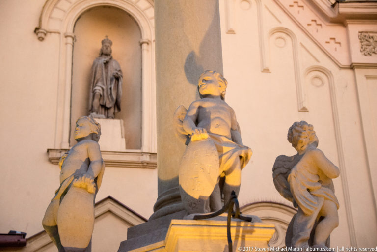 Three stone putti around a devotional monument, with a standing saint in a niche on the church façade behind.