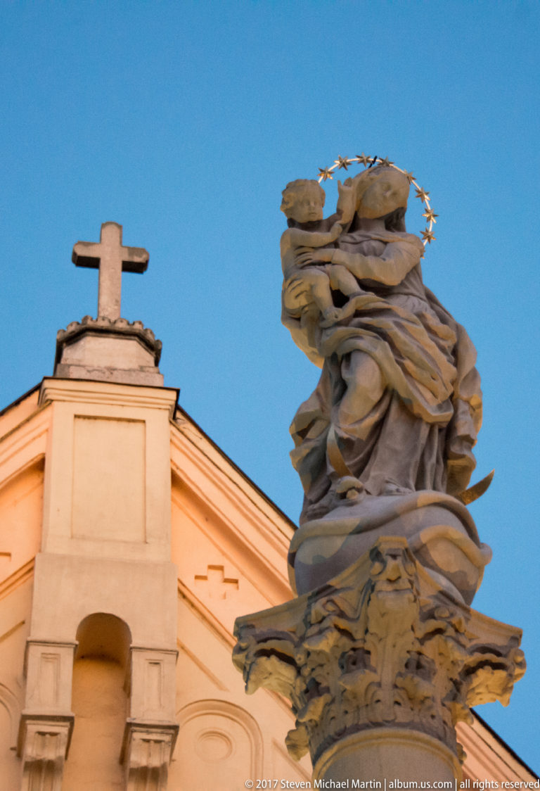 Crowned Virgin and Child stone sculpture with star halo beside a church gable topped by a cross at dusk.