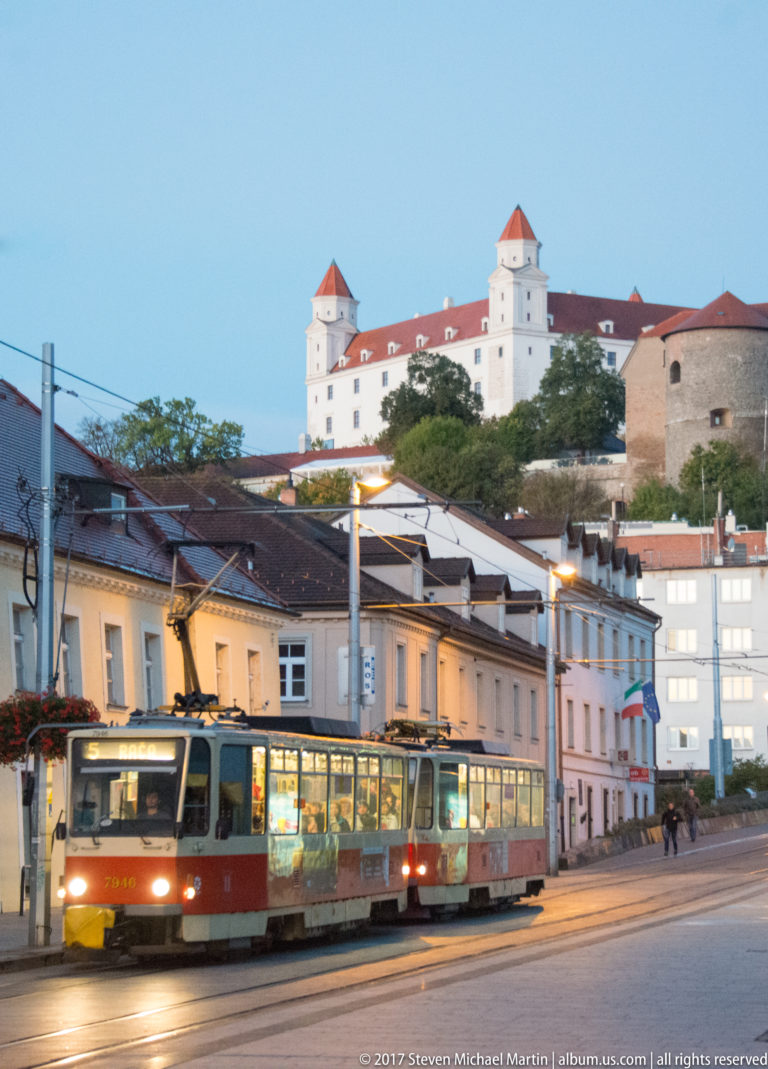 Red-and-cream tram on a Bratislava street at dusk, with Bratislava Castle rising on the hill above.