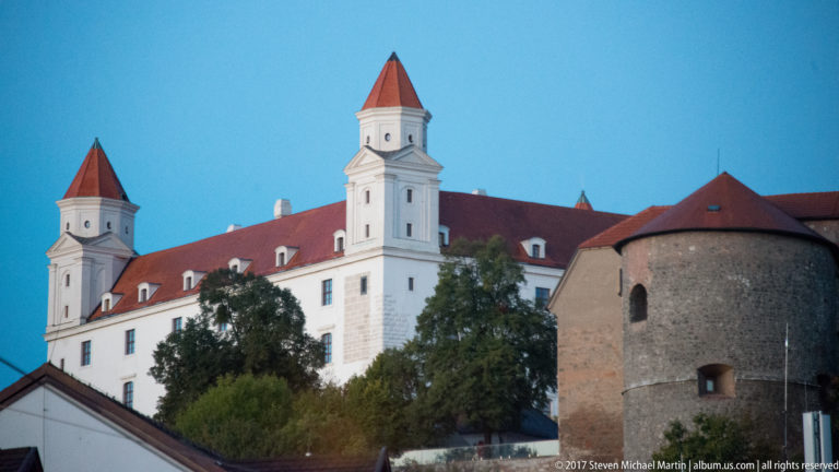 Bratislava Castle at dusk with white walls and red roofs rising behind a round stone defensive tower.