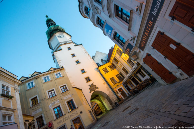 Tilted evening view up a Bratislava Old Town street toward Michael’s Gate and its green copper-roofed tower.