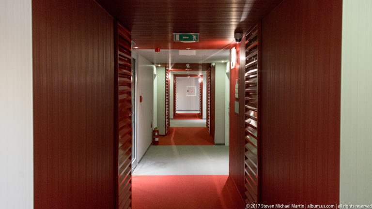 Long hotel corridor with dark wood panels, red carpet, repeating doorways, and bright opening at the far end.