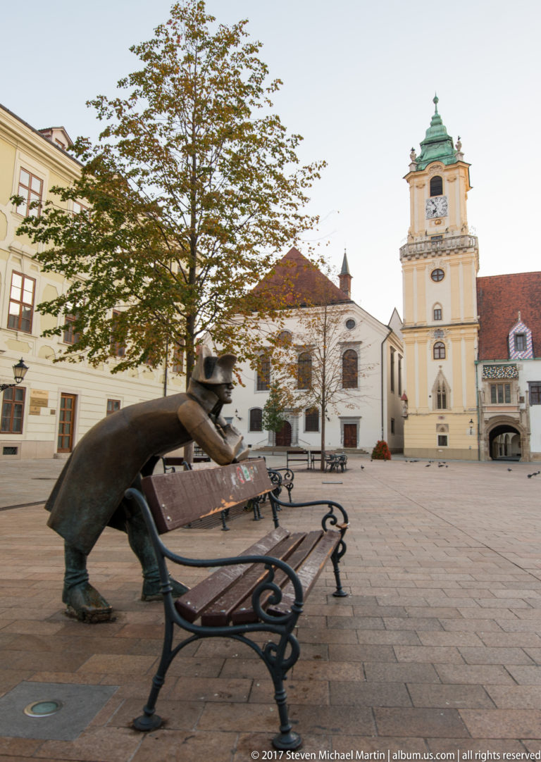 Profile view of the bronze Napoleon’s Army Soldier leaning over a bench, with a church tower and square behind.