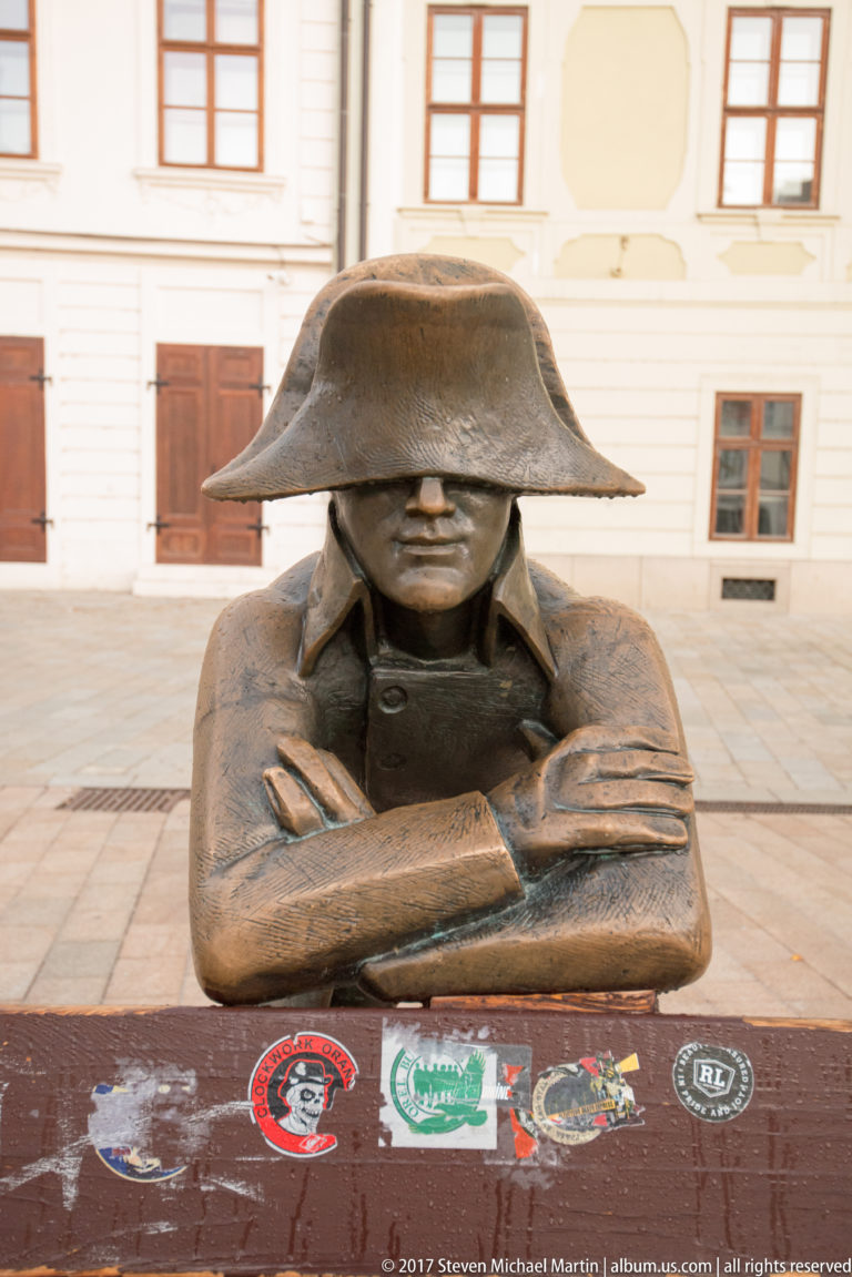 Close frontal view of the bronze Napoleon’s Army Soldier with bicorne hat and crossed arms resting on a bench.