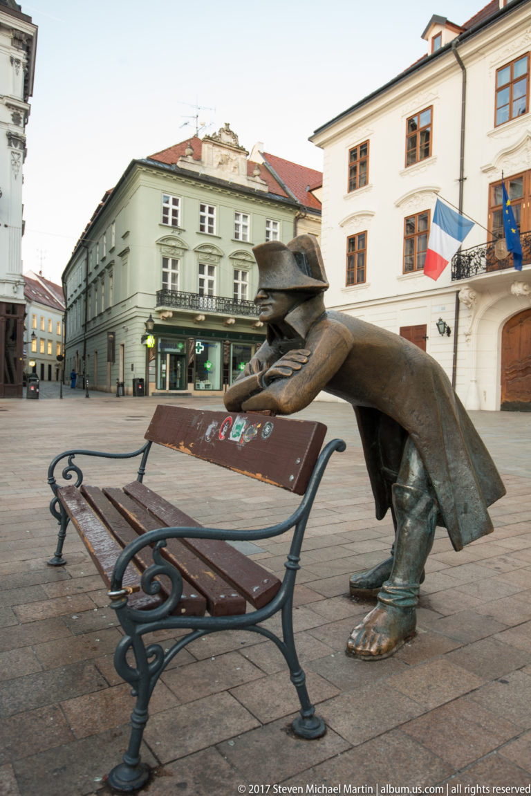Bronze Napoleon’s Army Soldier statue in a bicorne hat leaning over the back of a bench in Bratislava.