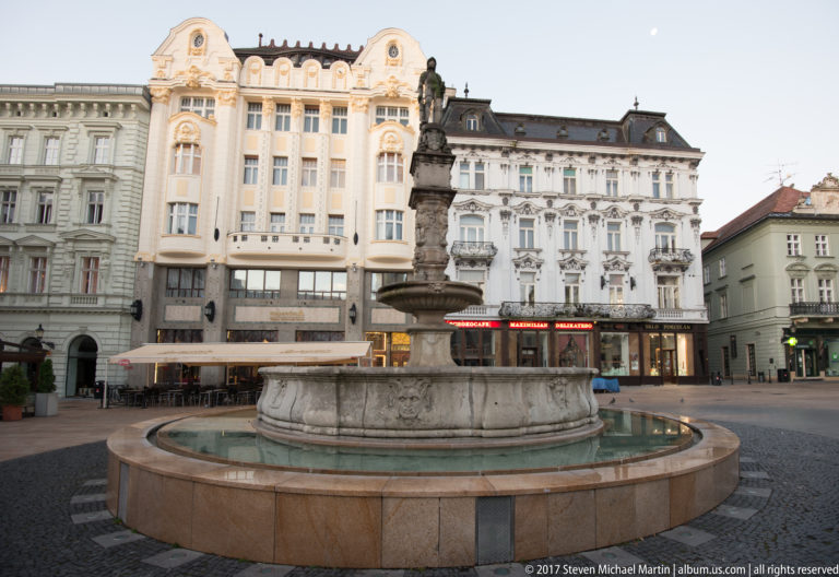 Maximilian Fountain centered in Bratislava Main Square, surrounded by ornate historic buildings and a broad paved plaza.