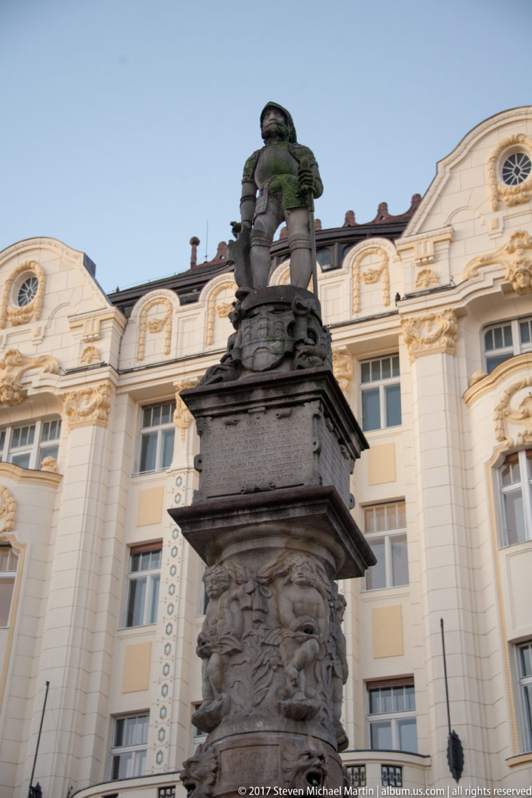 Stone statue and carved pedestal of the Maximilian Fountain seen from below against ornate buildings in Bratislava Main Square.