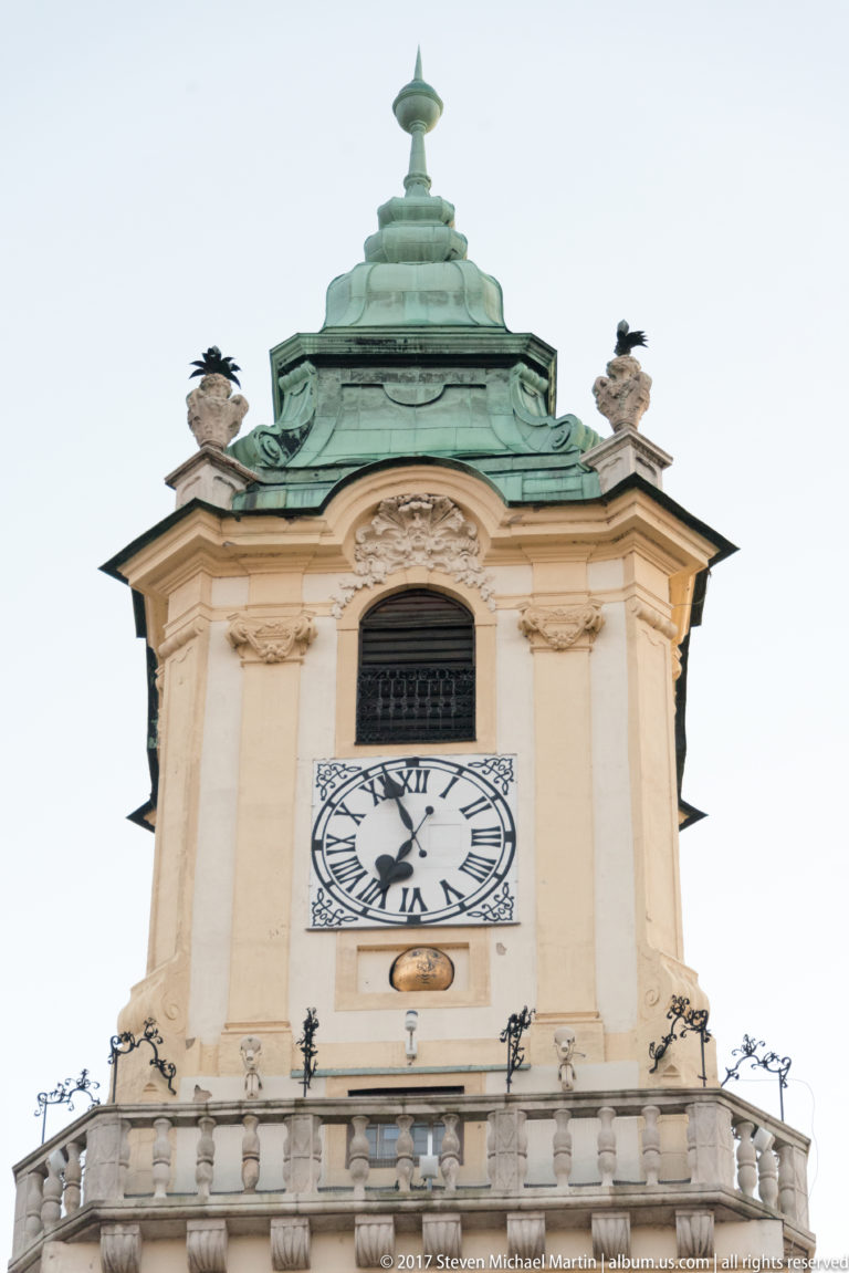 Baroque clock tower with Roman-numeral clock face, balustrade, small statues, and green patinated roof.