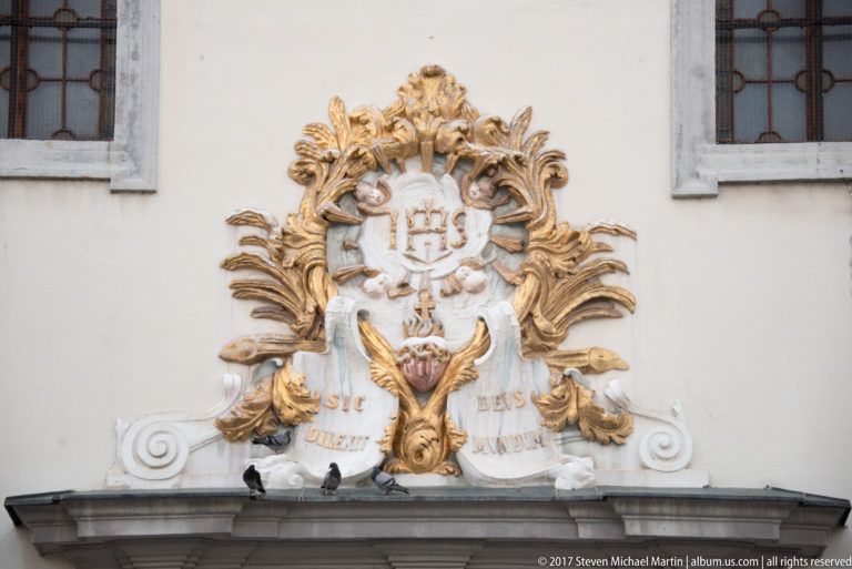 White and gold stucco cartouche with IHS monogram, foliage, and a gilded spread-winged bird on a façade.