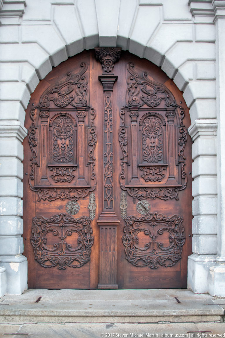 Ornately carved brown wooden double doors set within a pale rusticated stone arch in Bratislava.