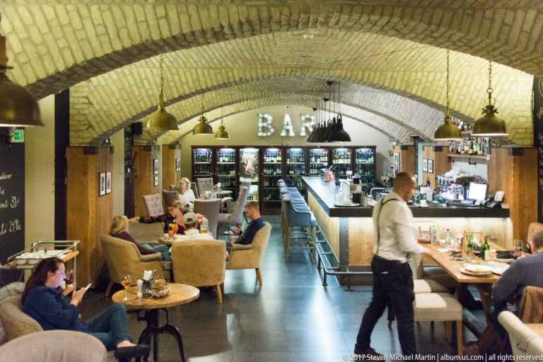 Brick-vaulted restaurant and bar with diners, waiter, hanging lamps, long counter, and refrigerated displays.