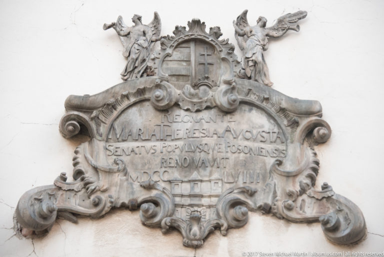 Elaborate carved stone cartouche with Latin inscription, scrollwork, and winged figures on a pale wall.