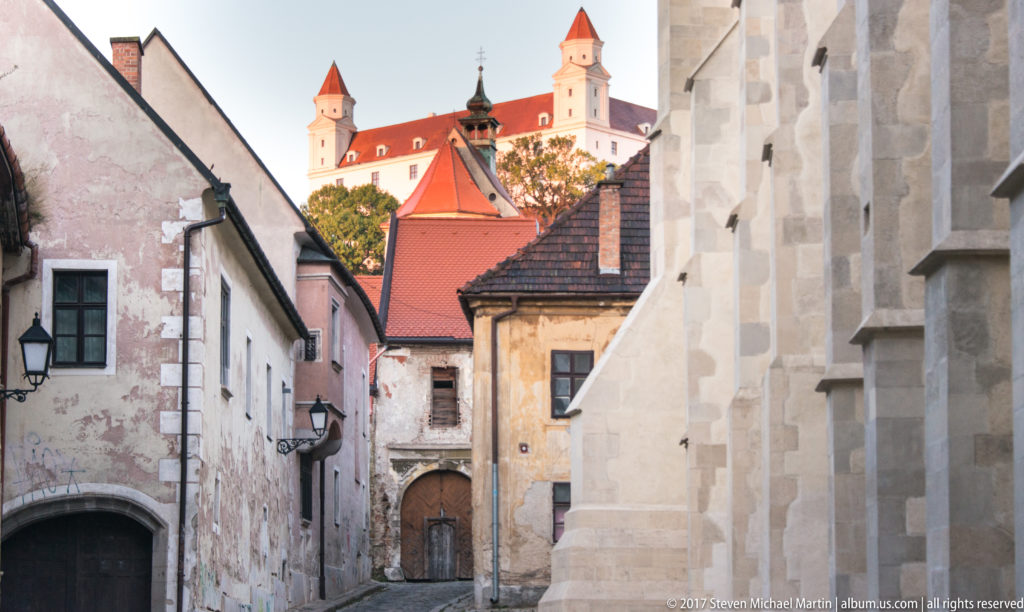 Bratislava Castle with white walls and red roofs seen above a narrow lane of older masonry buildings.