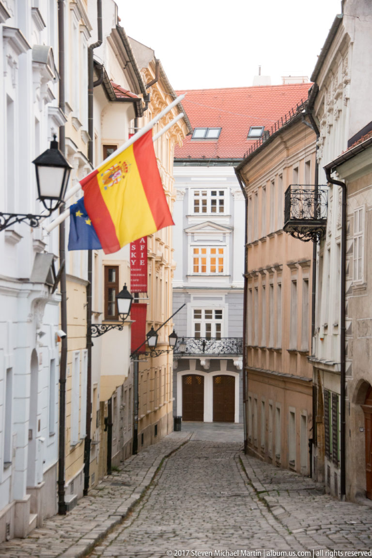 Cobbled Bratislava lane between historic buildings, with Spanish and European Union flags hanging above the street.