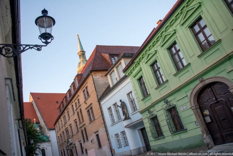 Narrow Bratislava Old Town street lined with pastel historic buildings and steep roofs, with a church spire rising in the distance.