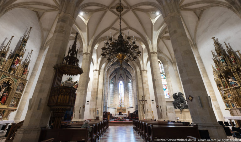 Central aisle of a Gothic church with ribbed vaults, chandelier, side altarpieces, pews, and distant sanctuary.