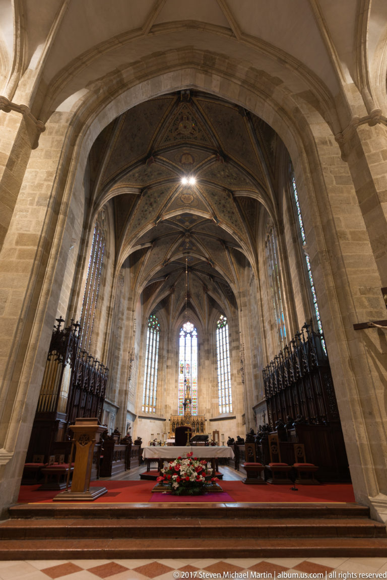 Gothic church nave looking toward a bright sanctuary with ribbed vaults, choir stalls, altar, and tall windows.