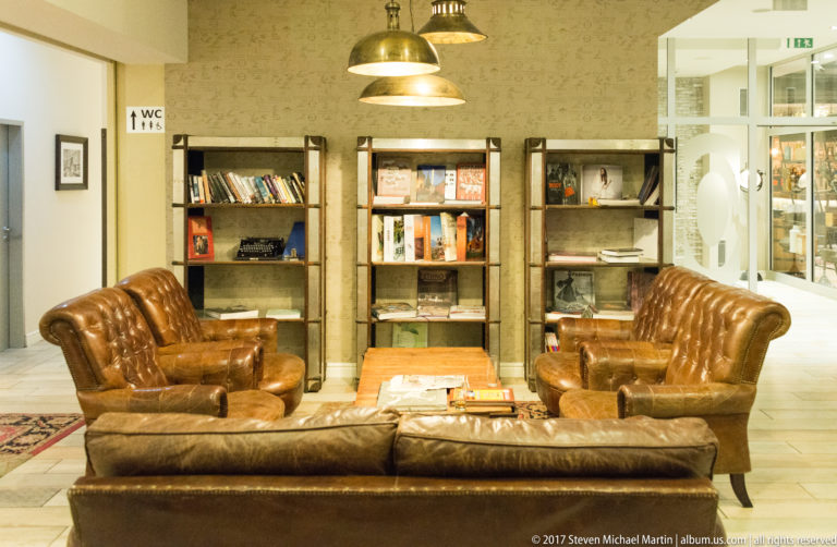 Brown leather armchairs and sofa in a library-like hotel lounge with shelves, display cabinets, and industrial pendant lights.