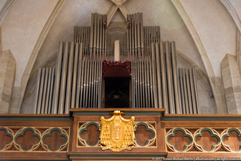 Tall church organ pipes above a decorated gallery with gilded crest and Gothic ribbed vaulting.