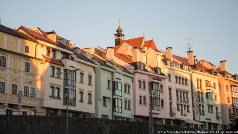 Pastel historic buildings with red roofs, dormers, chimneys, and a small tower in warm evening light.