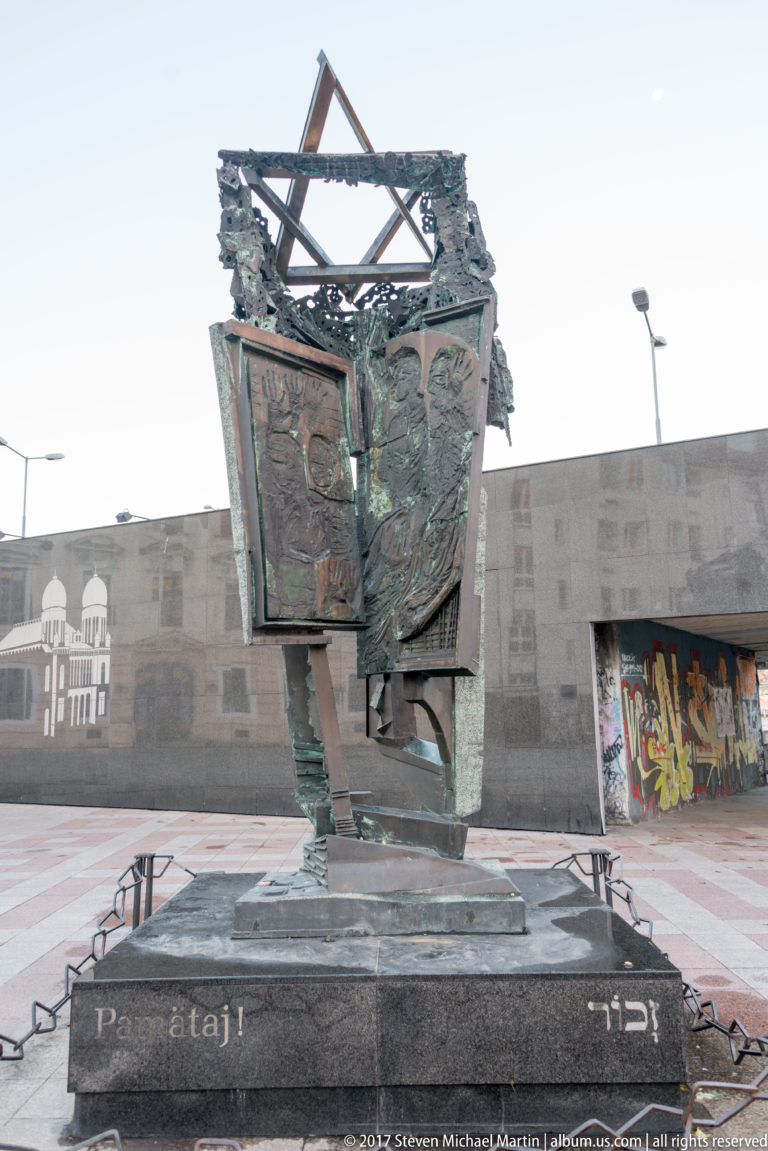 Angular bronze memorial with relief panels and broken open top on a stone plinth in a paved plaza.