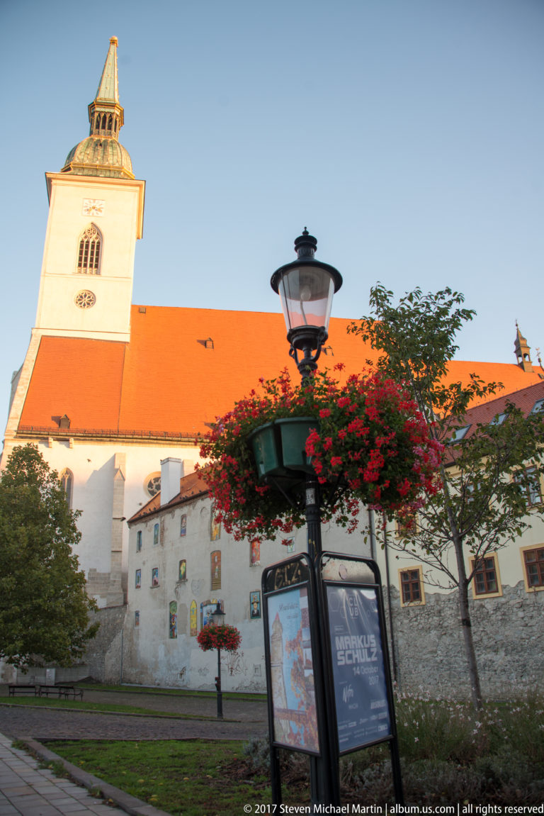 White clock tower and red-roofed historic church complex behind flowers and a streetlamp in Bratislava.