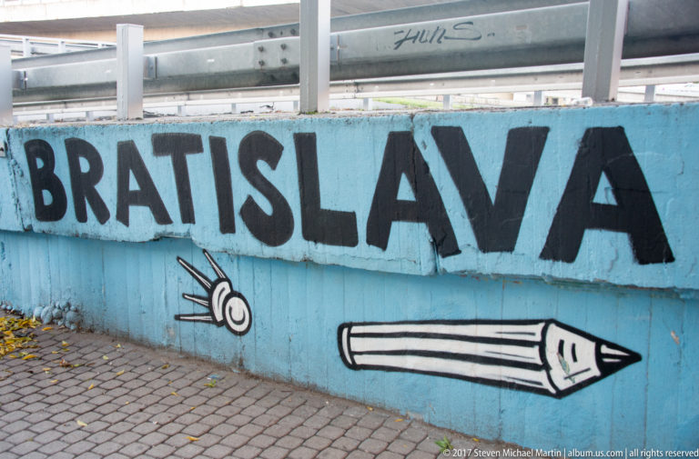 Large black “BRATISLAVA” lettering with snail and pencil drawings on a blue concrete wall.