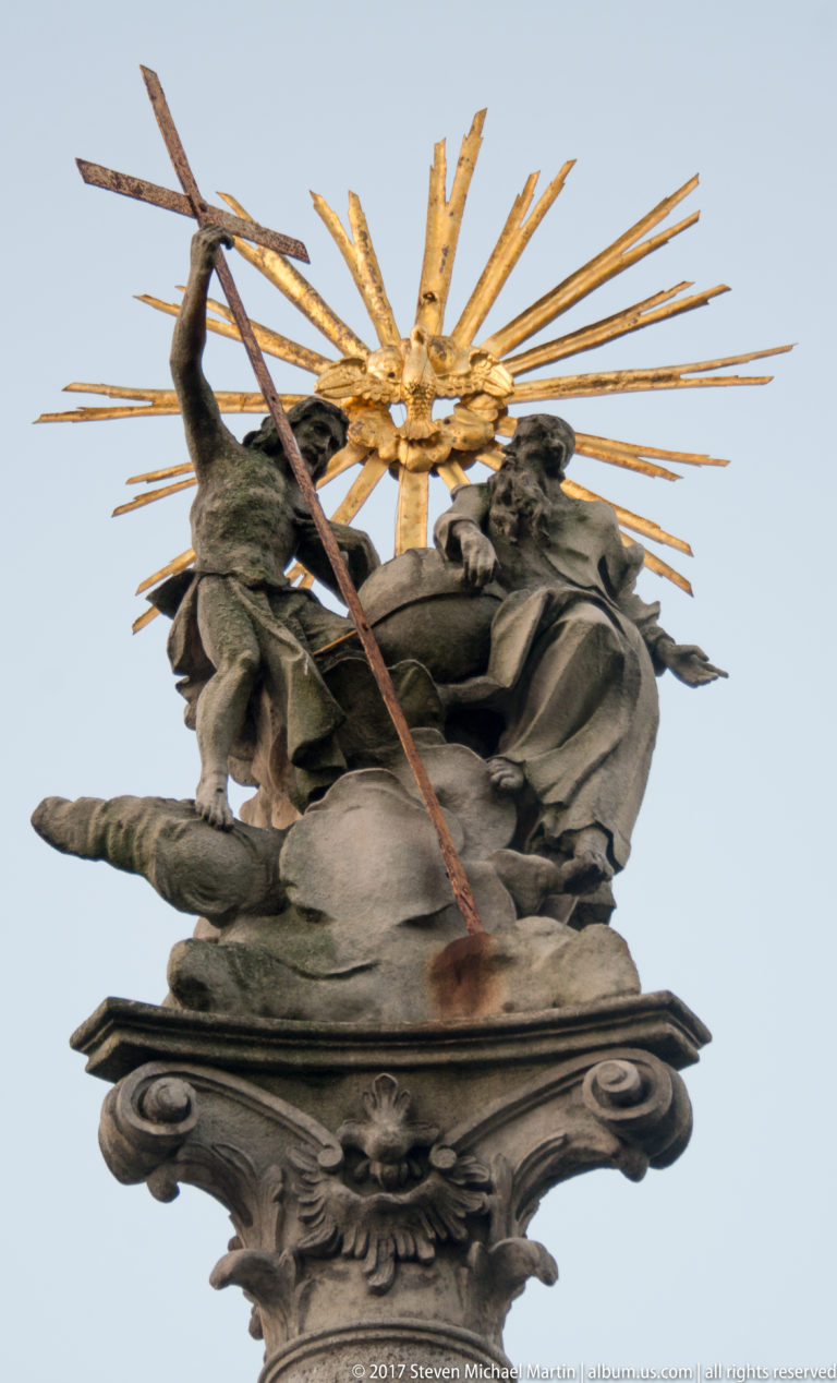 Baroque stone figure group backed by a large gilded sunburst, viewed against the sky.