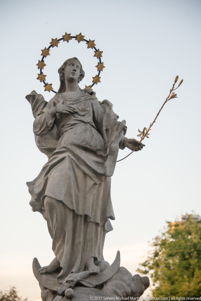 Stone female saint with starry halo, flowing robes, and a flowering branch, standing on a crescent-shaped base.
