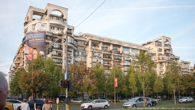 Curved multi-story apartment block above a tree-lined Bucharest intersection with cars, pedestrians, traffic lights, and overhead wires.