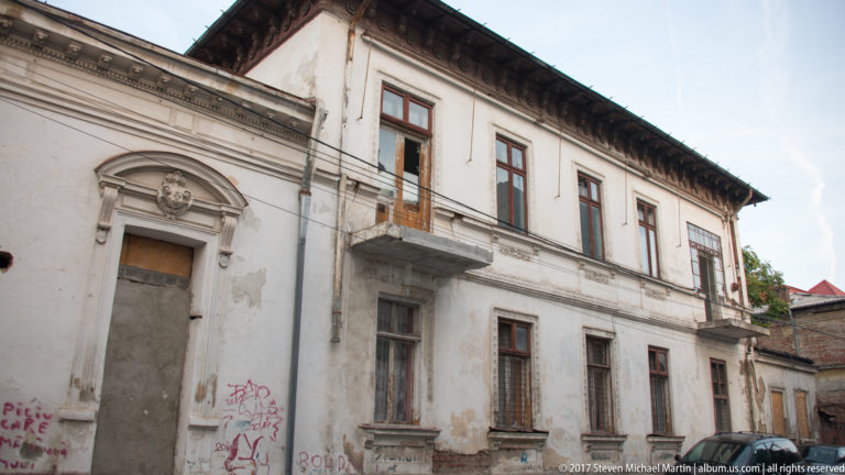 Weathered two-story Bucharest house with tall windows, classical moldings, small balcony, graffiti, and patched stucco.