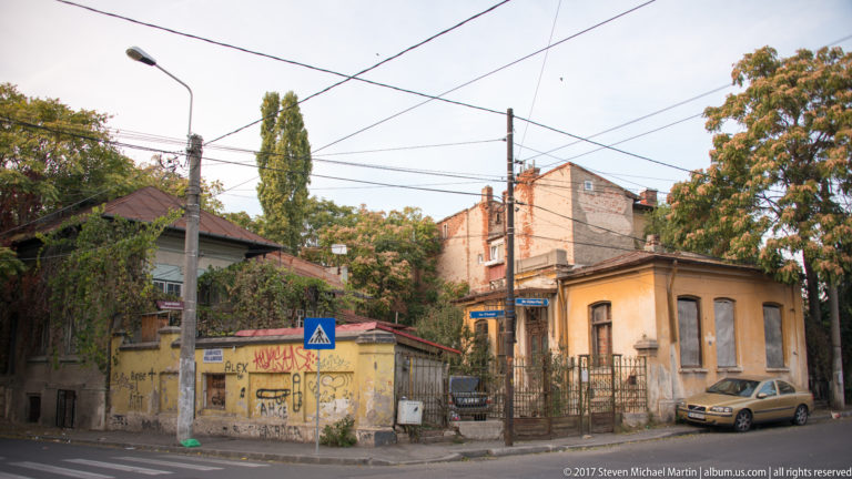 Older low-rise houses with worn plaster, graffiti, metal gates, trees, parked car, and overhead wires at an intersection.