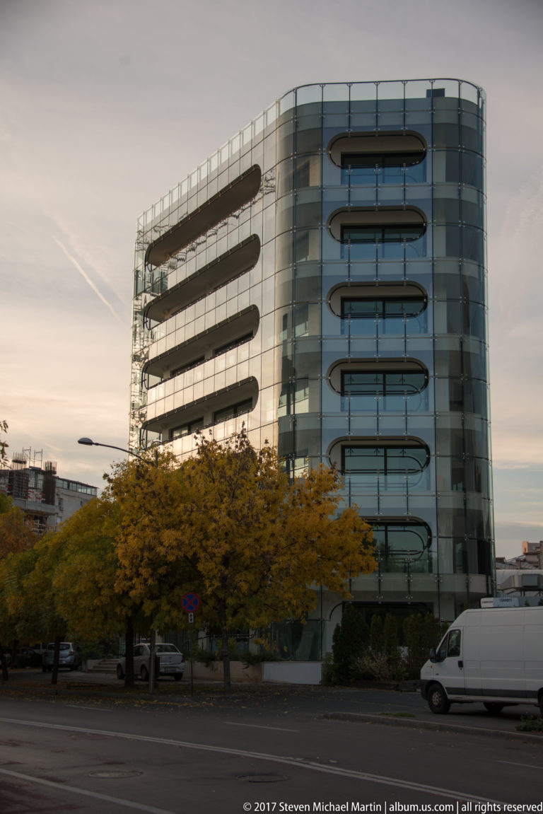 Curved modern glass office building with horizontal balcony bands, trees, and a white van along the street.