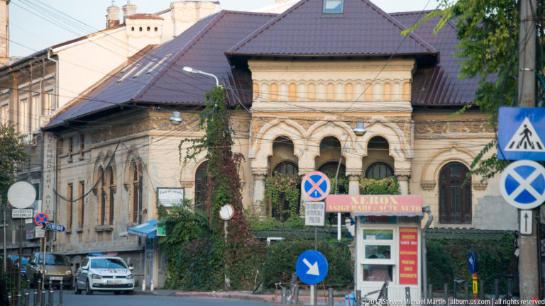 Historic Bucharest building with paired round-arched windows, tiled roof, decorative masonry, ivy, and street signs.
