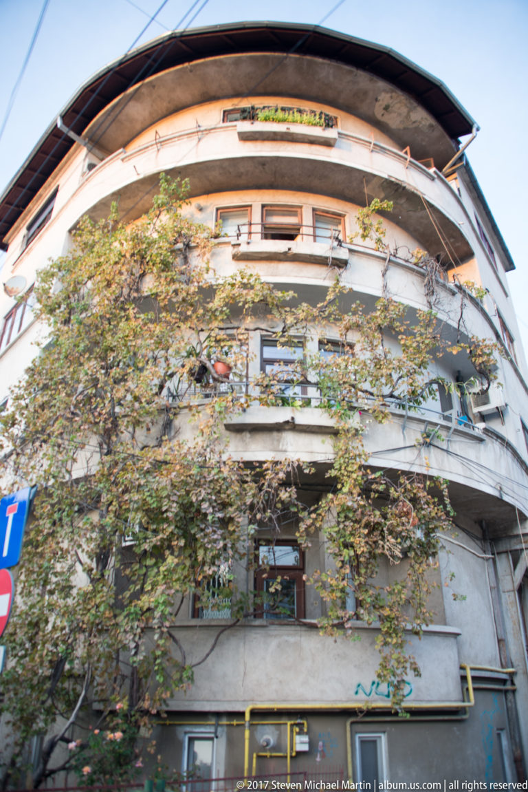 Rounded-corner Bucharest apartment block with continuous balconies, trees, street signs, and utility lines.