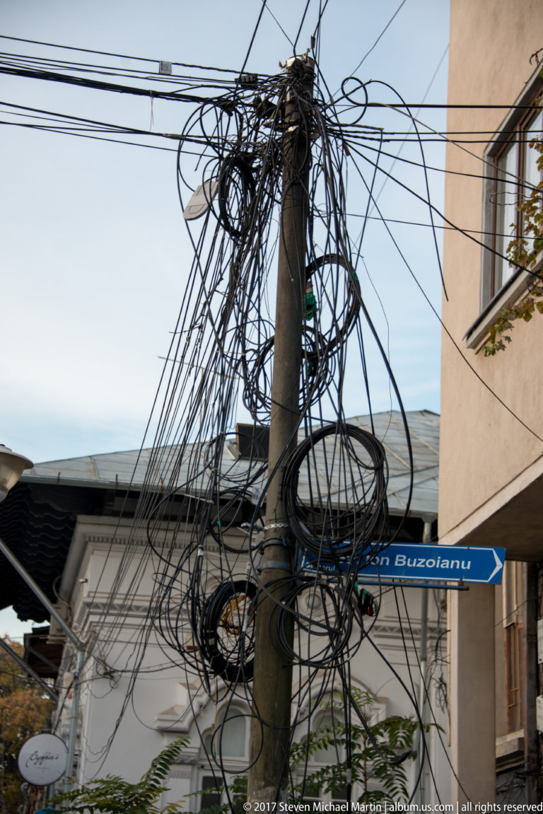 Utility pole wrapped in dense loops of overhead cable between buildings, with a blue directional street sign behind.