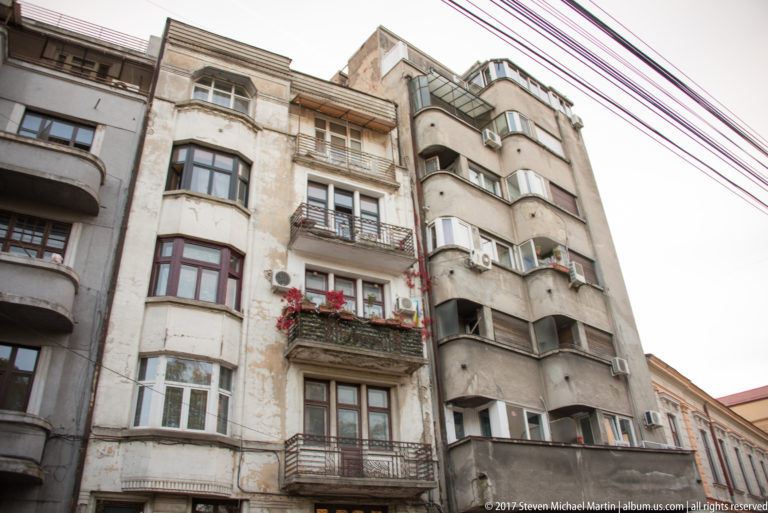 Weathered Bucharest apartment building with rounded bays, balconies, flower boxes, patched stucco, and overhead cables.