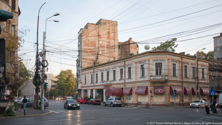 Two-story pale corner building at a Bucharest intersection, with awnings, cars, pedestrians, and dense overhead wiring.