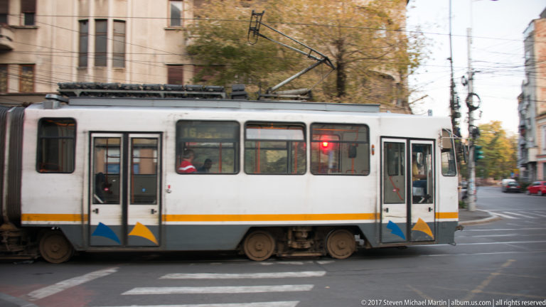White Bucharest tram crossing a city intersection under overhead wires, with passengers visible through the windows.